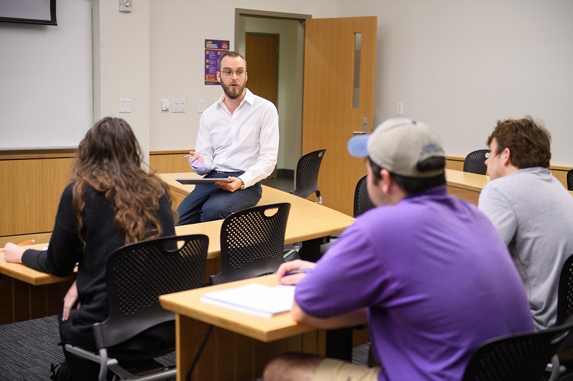 Teacher sits on desk while lecturing as students look on