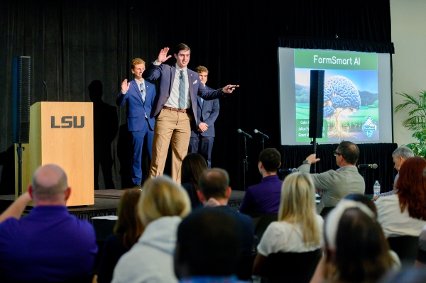 Three men present on a stage. One is raising his hand and pointing to large display screen that says FarmSmart AI.